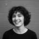 Smiling young woman with short, curly dark hair, wearing a dark top, against a soft gray background.